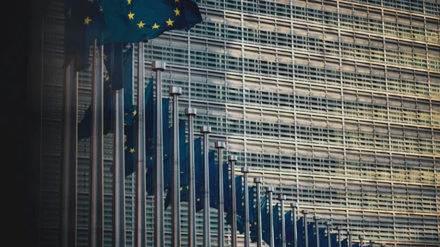 EU-flaggor på flaggstänger i rad framför en stor skyskrapa.