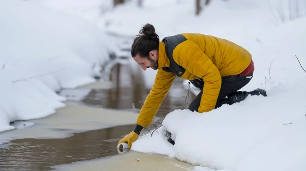 A man leans over a river surrounded by snow and scoops up some water